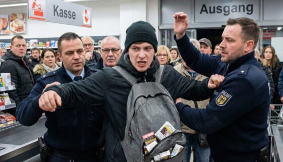 Gewalttätiger Kampf am Supermarktausgang: Ein Mann schlägt einen Beamten; Sicherheitsleute halten ihn fest, der Rucksack quillt vor Zigarettenschachteln.