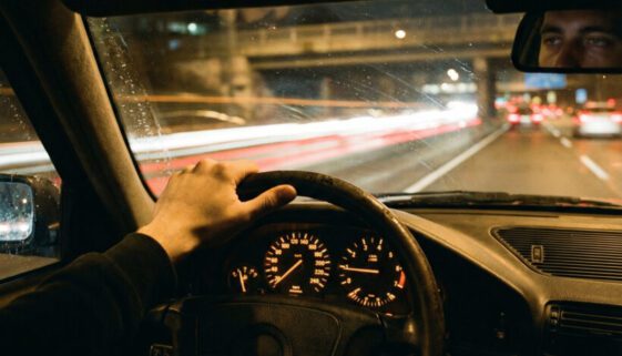 Die lockere Hand eines übermüdeten Mannes am Lenkrad eines dunklen Wagens, umgeben von verschwommenen, grellen Autobahnlichtern.