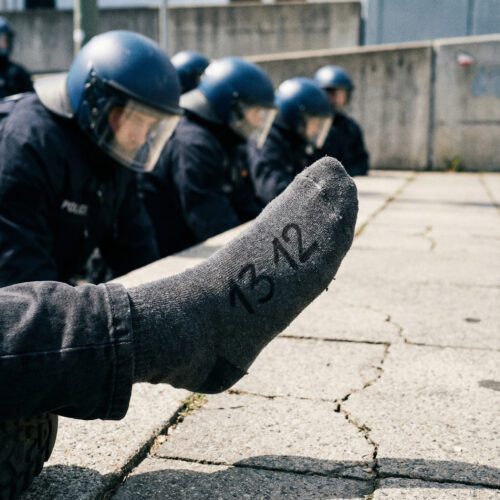 Nahaufnahme der Zahl „13 12“ auf der angehobenen Socke eines Demonstranten, konfrontativ auf uniformierte Polizeibeamte gerichtet.