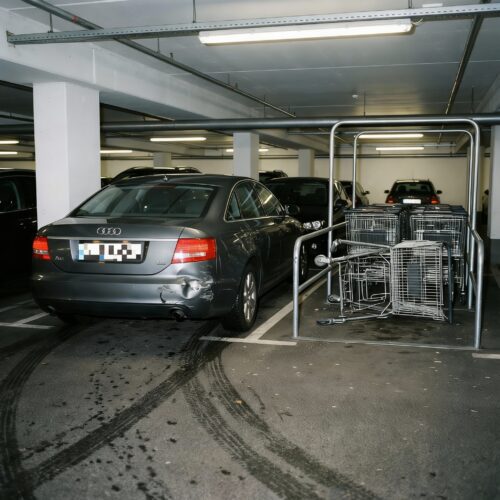 Ein moderner PKW mit Kollisionsschaden am Heck fährt eilig am verbogenen Einkaufswagen-Gestell vorbei.