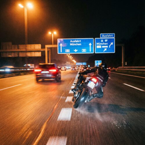 Ein rasant geneigtes Motorrad in extremer Schräglage rast bei Nacht knapp an den Rücklichtern anderer Fahrzeuge vorbei.