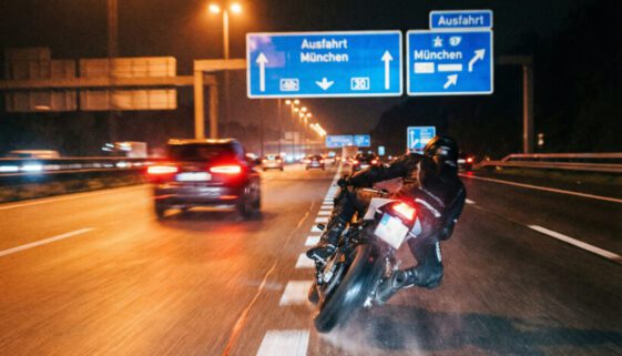 Ein rasant geneigtes Motorrad in extremer Schräglage rast bei Nacht knapp an den Rücklichtern anderer Fahrzeuge vorbei.