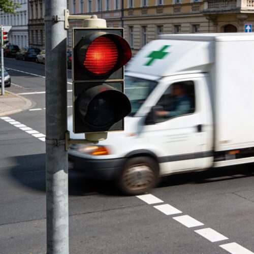 Ein weißes Apotheken-Lieferfahrzeug fährt eilig bei rot leuchtender Ampel über die Haltelinie einer Kreuzung.