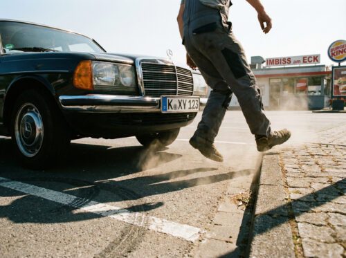 Ein dunkles Auto nähert sich aggressiv den Beinen eines Mannes, der knapp auf eine Steineinfassung zur Seite springt.
