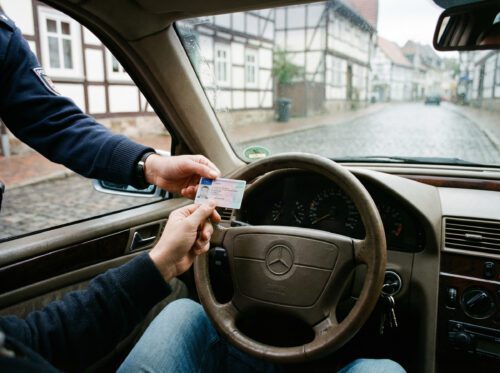 Ein Fahrer am Lenkrad hält zögerlich seinen EU-Führerschein, während eine polizeiliche Hand bestimmt danach greift.