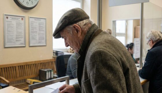 Ein älterer Mann schiebt einen handgeschriebenen Zettel unter die Glasscheibe eines kargen Behördenschalters.