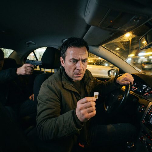 Eine SIM-Karte in einer verkrampften Hand, während im dunklen Auto ein bedrohlicher Pistolenlauf auf den Mann zielt.