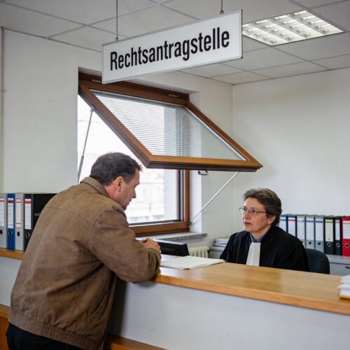 Ein laienhafter Mann spricht am Schalter der Rechtsantragstelle eines Gerichts mit einem Justizbeamten.