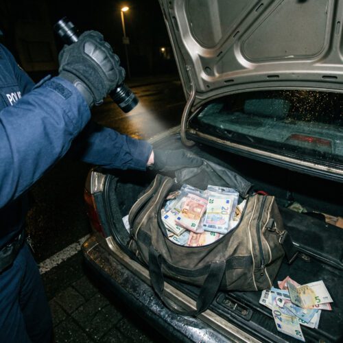 Eine behandschuhte Hand greift im Lichtkegel nach chaotisch gebündelten Geldscheinen in einer Sporttasche.