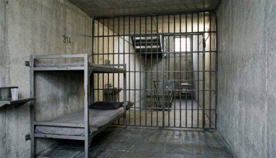 Karge deutsche Gefängniszelle mit massivem Stahlbett, grauen Betonwänden und dicken Gitterstäben im Vordergrund.