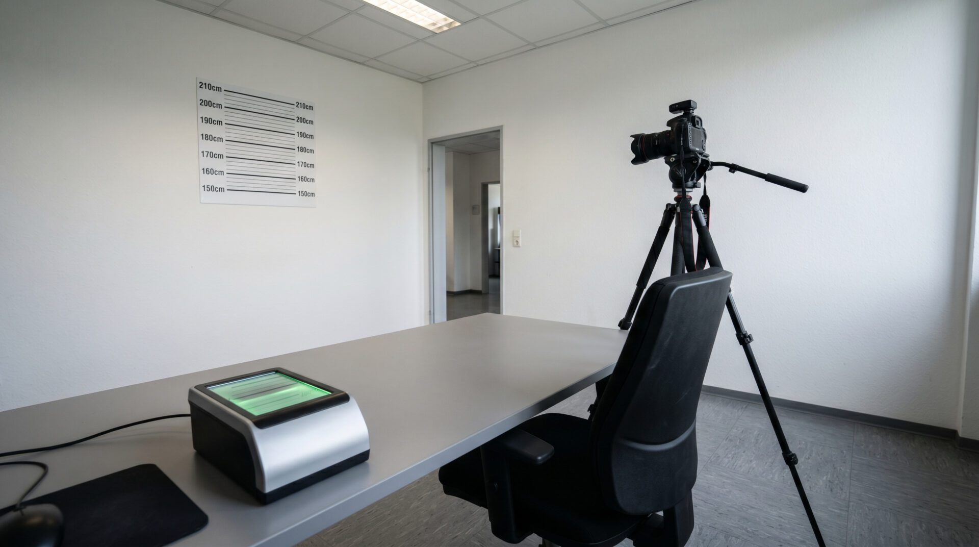 Leerer Stuhl vor einer Fotowand mit Größenskala und einem digitalen Fingerabdruck-Scanner in einer Polizeidienststelle.