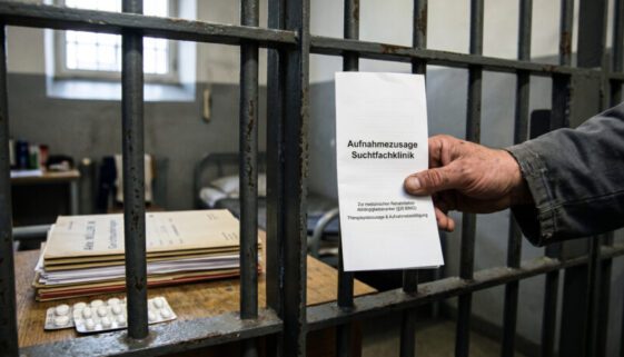 Hand eines Inhaftierten hält Klinik-Broschüre hinter Gefängnisgittern neben Gerichtsdokumenten.